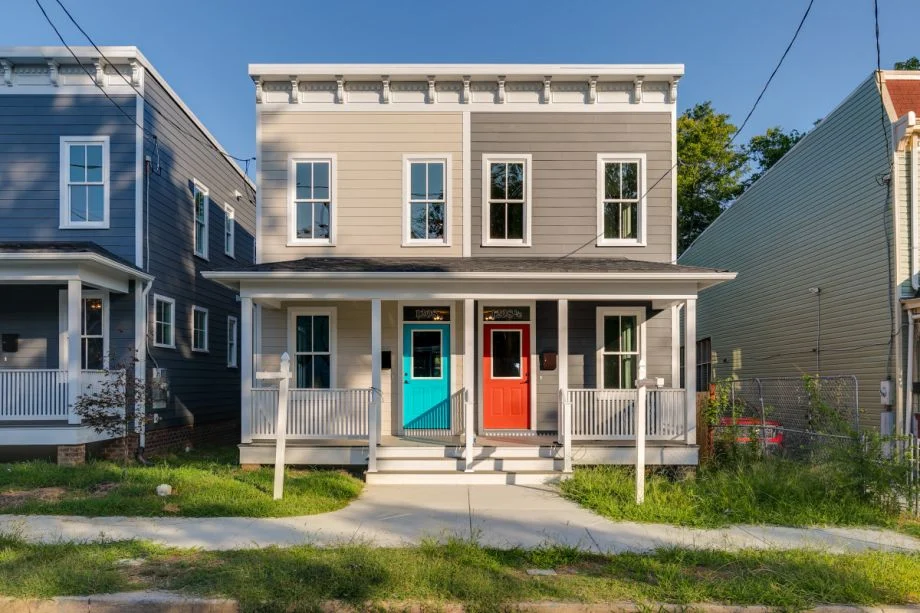 Two-story duplex with gray siding, white trim, a front porch, teal door on the left unit, and orange door on the right unit. The house is flanked by similar homes and has a small, grassy front yard.