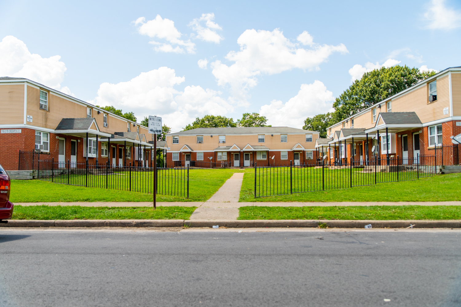 A wide view of a residential apartment complex with two-story brick and beige buildings, a green lawn in the center, and a sidewalk leading from the street through a gated fence.