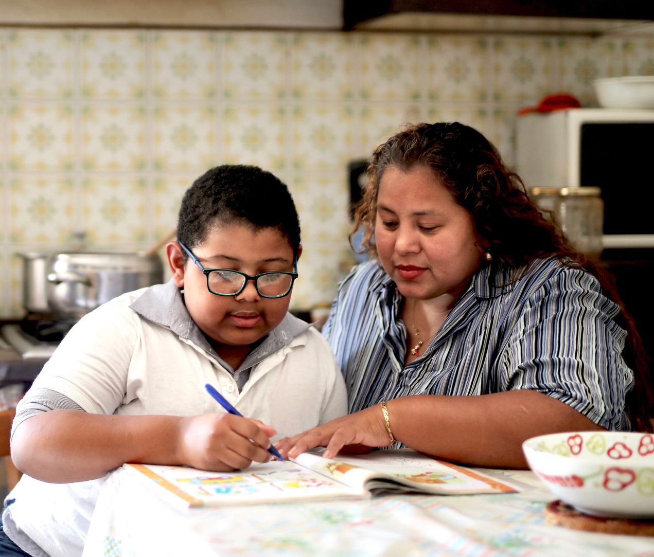 A woman helps a young boy with glasses as he writes in a workbook at a kitchen table. They both focus on the activity, with kitchen items and a microwave visible in the background.