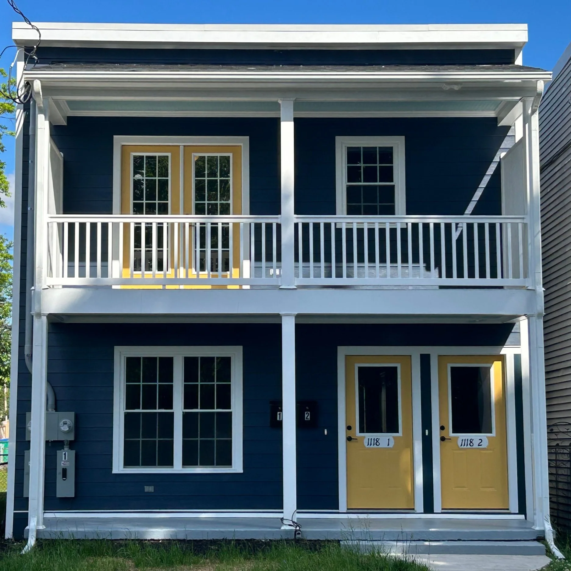 A two-story blue house with white trim, two yellow-front doors labeled 1118 1 and 1118 2, and a second-floor balcony with matching yellow French doors. Grass lawn in front and blue sky above.