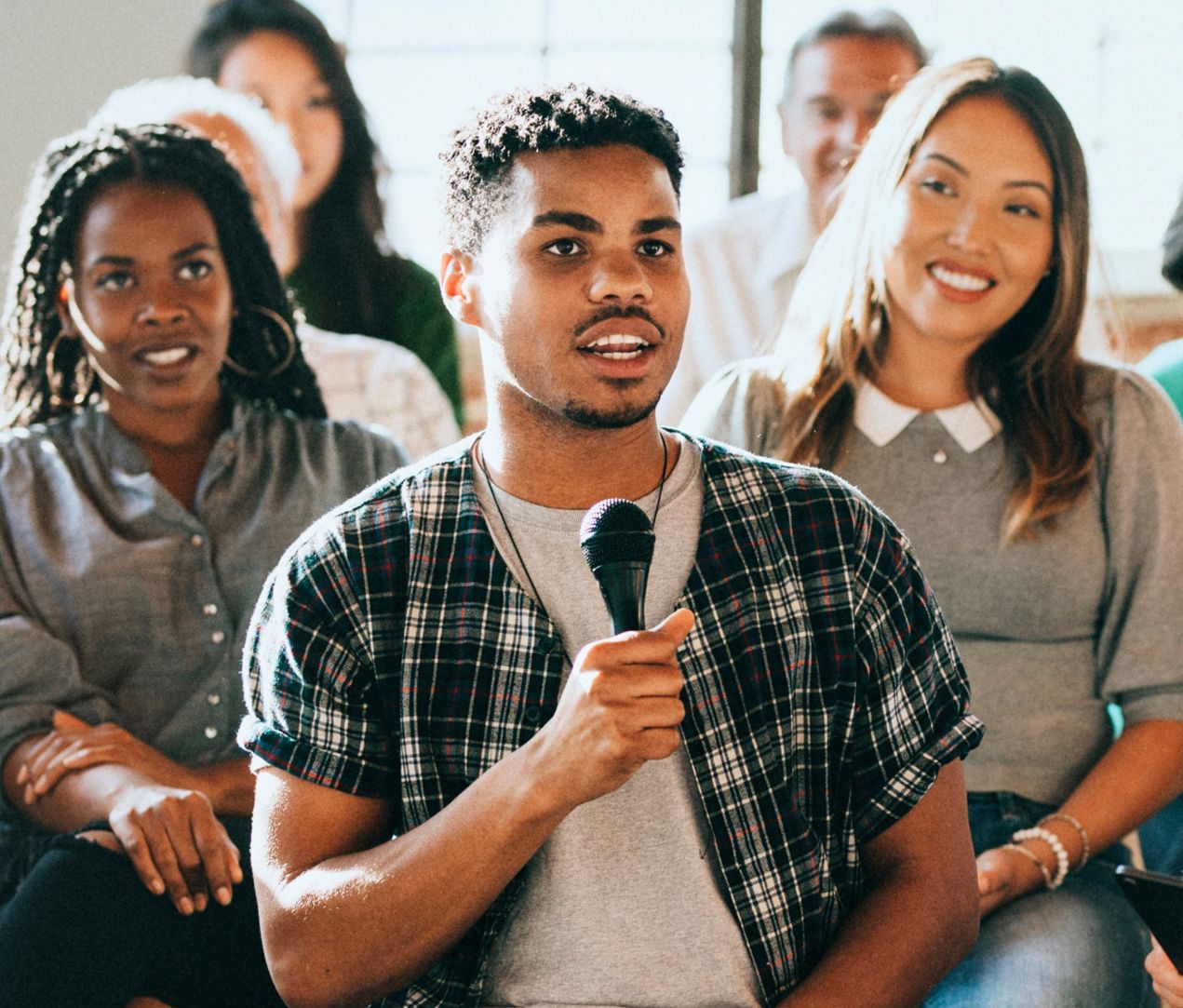 A young man holds a microphone and speaks in a group setting, with several people seated and smiling behind him, suggesting a friendly and engaged audience.