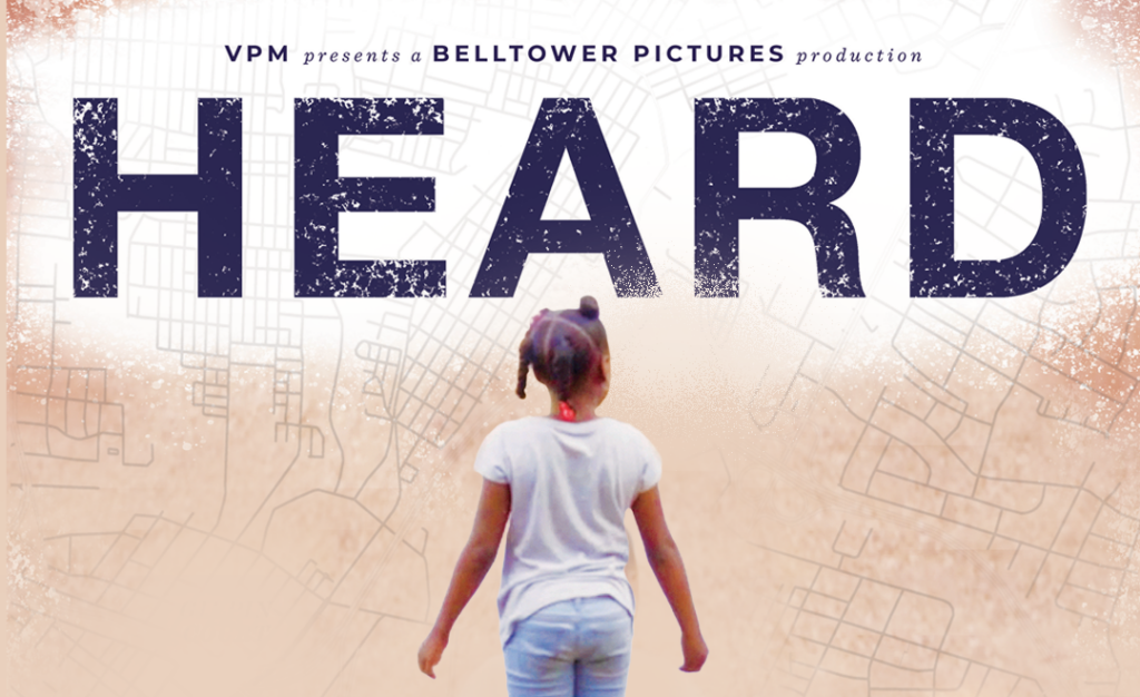 A young girl with two hair buns, wearing a white shirt and jeans, stands with her back to the camera facing large bold text that reads HEARD on a light, abstract background.