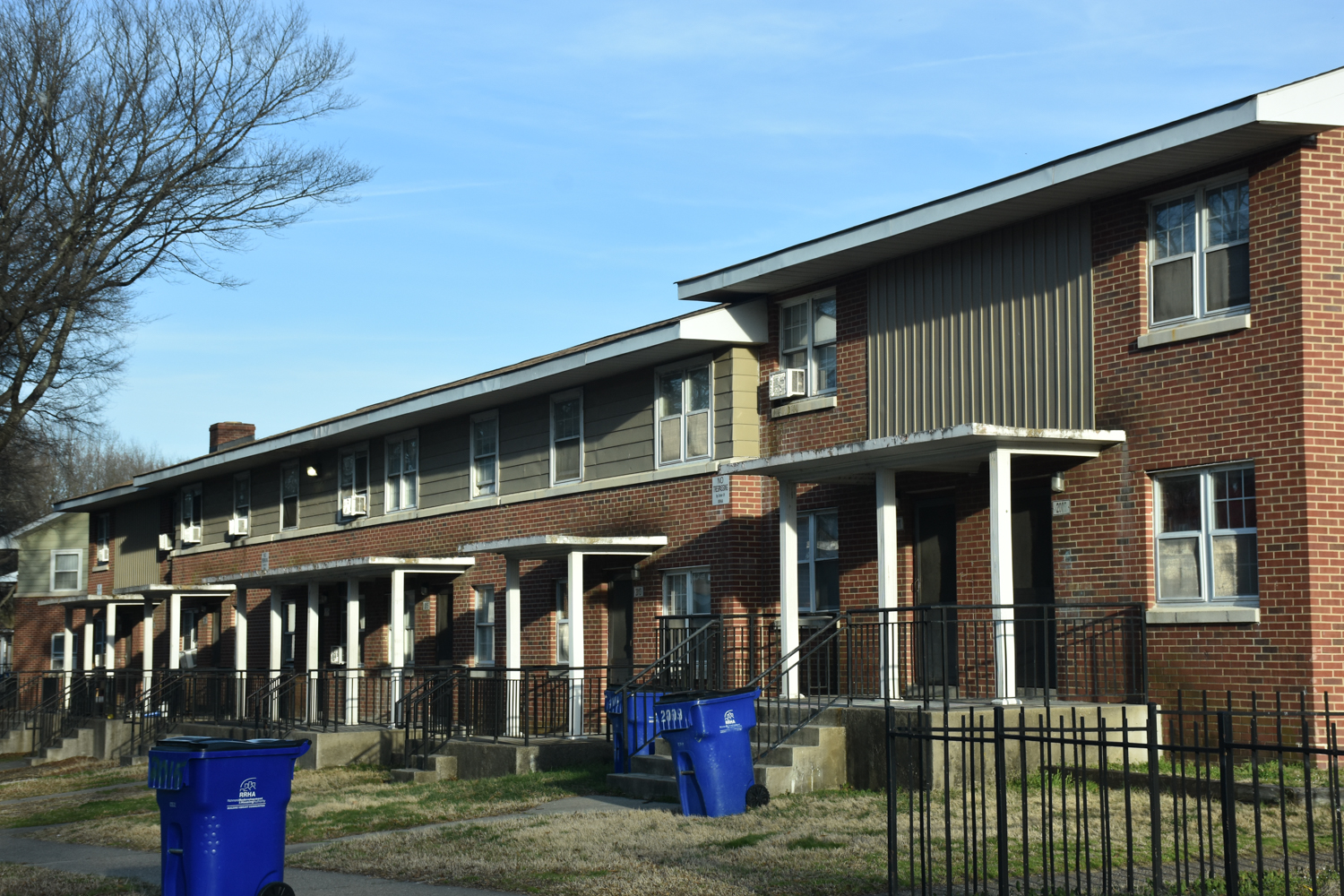 A row of two-story brick apartment buildings with small covered porches and black railings. Blue recycling bins are on the sidewalk, and bare trees stand nearby under a blue sky.