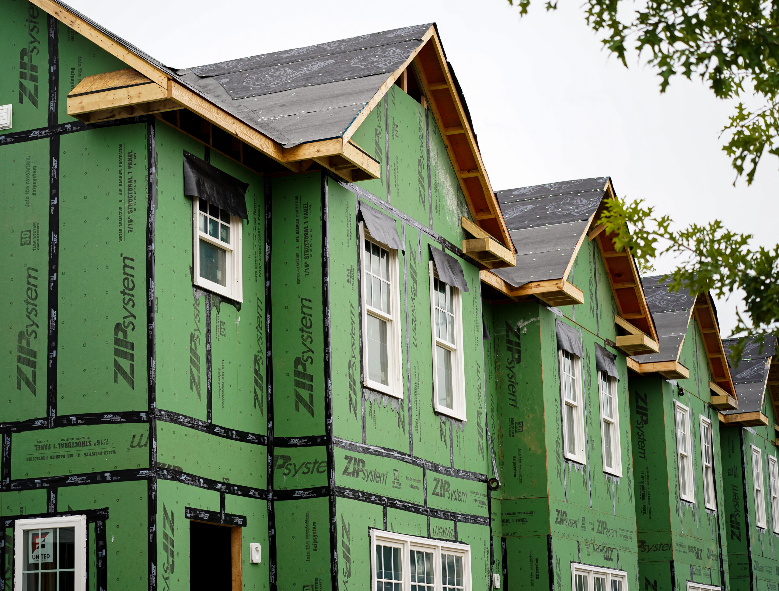 A row of multi-story townhouses under construction, covered in green sheathing labeled ZIP System, with visible windows and unfinished roofs against a cloudy sky.