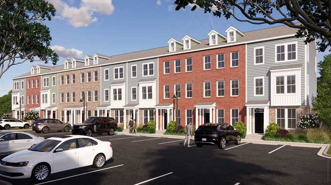 A row of modern, three-story townhouses with brick and siding exteriors faces a parking lot with several parked cars under a blue sky with scattered clouds. Trees frame the scene.