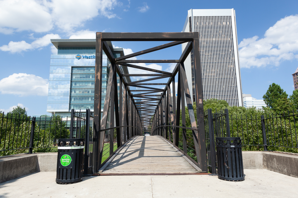 A metal pedestrian bridge with a wooden walkway leads towards modern office buildings. Trees and clouds are visible under a blue sky. Trash cans and a green sign flank the bridge entrance.