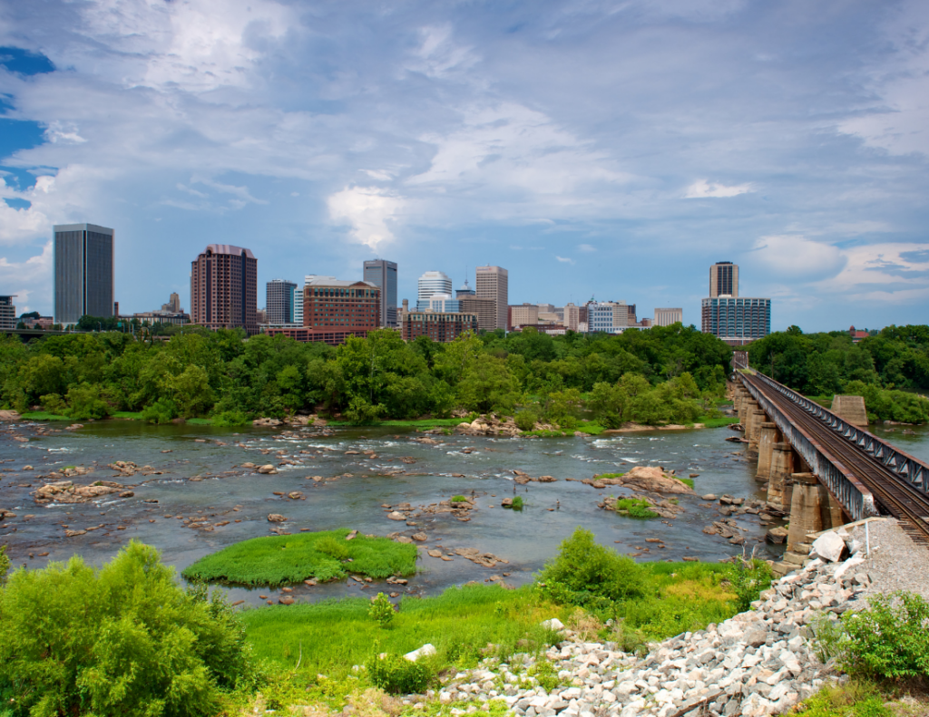 View of a city skyline with modern buildings behind a river, scattered rocks, and lush green trees. A railroad bridge crosses the river on the right under a partly cloudy blue sky.