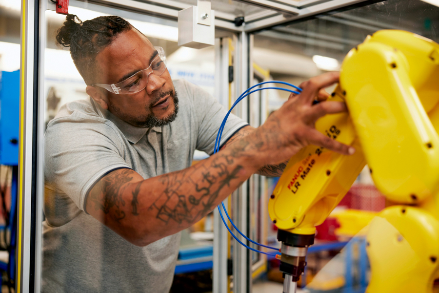 A man wearing safety glasses adjusts a yellow robotic arm inside a glass-enclosed workspace, focusing intently on his task. Blue wires and machinery are visible in the background.
