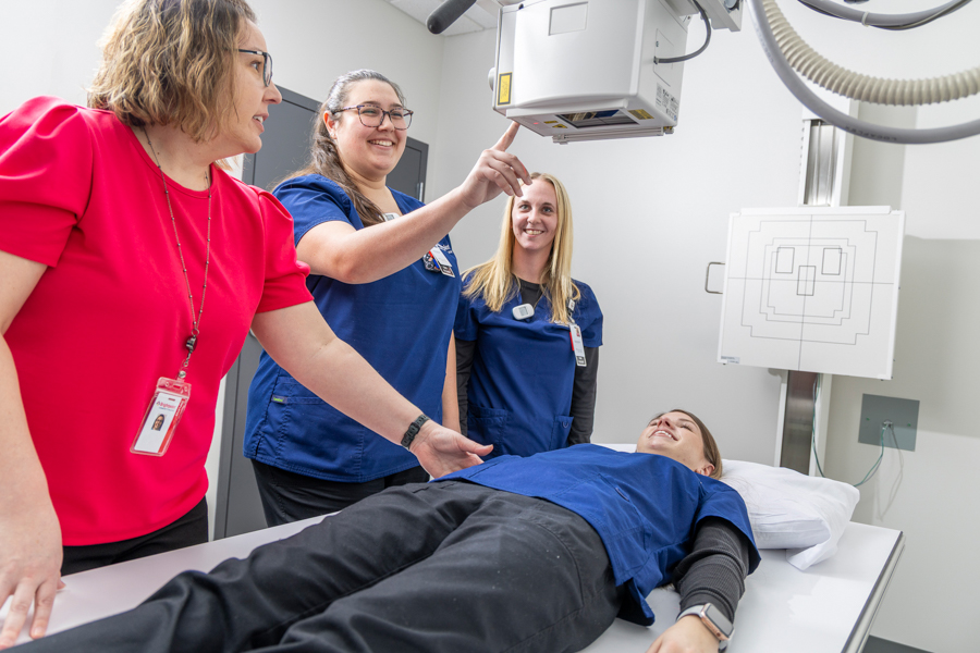 Four women in a medical imaging room; one woman lies on an exam table while the others, in scrubs, operate and discuss the X-ray machine overhead. They appear focused and collaborative.