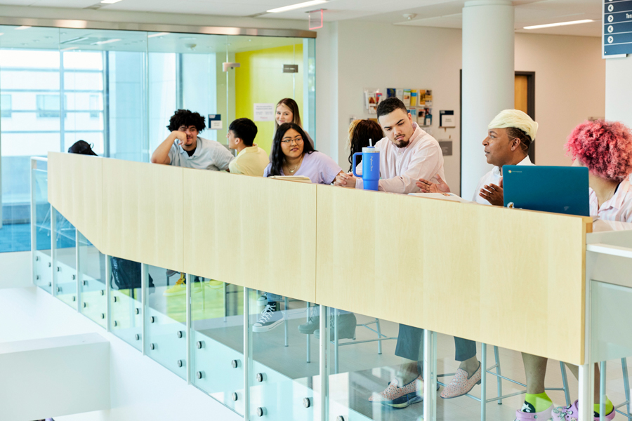 A group of diverse students sits along a railing in a modern indoor space, engaged in conversation and using laptops, with large windows in the background letting in natural light.
