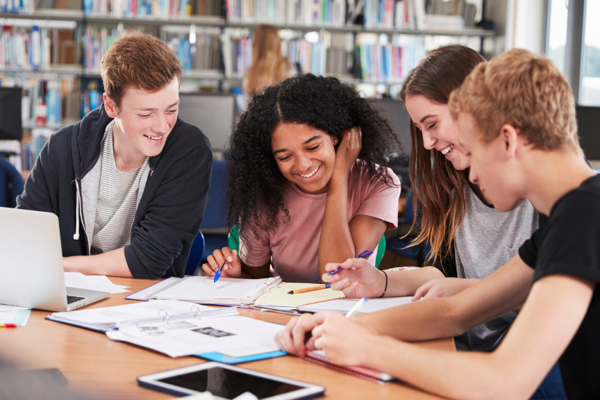 Four students sit together at a table in a library, smiling and working on schoolwork with notebooks, paper, and a laptop and tablet in front of them. Bookshelves are visible in the background.