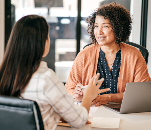 Two women sit at a desk having a conversation in an office setting. One woman faces the camera, smiling and listening attentively, while the other woman has her back to the camera and gestures with her hands.