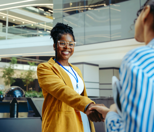 A woman wearing glasses and a mustard yellow blazer smiles and shakes hands with another person in a modern office lobby.