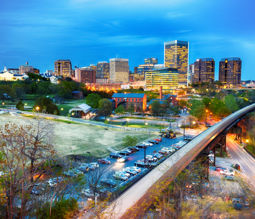 A city skyline at dusk with tall modern buildings lit up, a grassy park area, a red brick building, a busy street, and a parking lot in the foreground. Trees and a blue evening sky complete the scene.