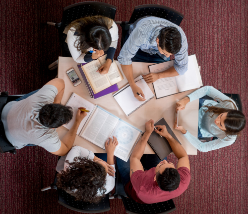 Six people sit around a table with books, notebooks, and a phone, working together and taking notes. The scene is viewed from above on a red carpeted floor.