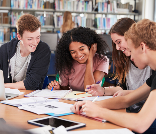 Four students sit together at a table in a library, smiling and working on notebooks and papers. Books, a tablet, and a laptop are scattered across the table, suggesting a collaborative study session.