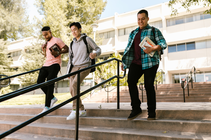 Three college students walk down outdoor steps in front of a school building, carrying books and backpacks. They are smiling and appear to be enjoying each others company on a sunny day.