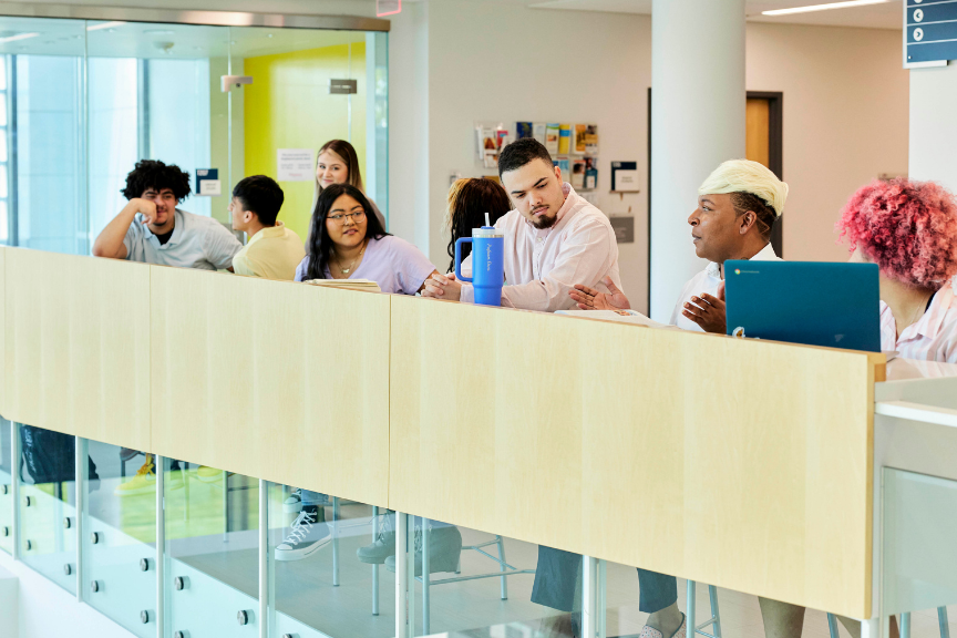 A diverse group of people sit and talk at a long wooden table in a bright, modern building, with glass walls and various materials visible in the background. Some are listening, while one person gestures as they speak.