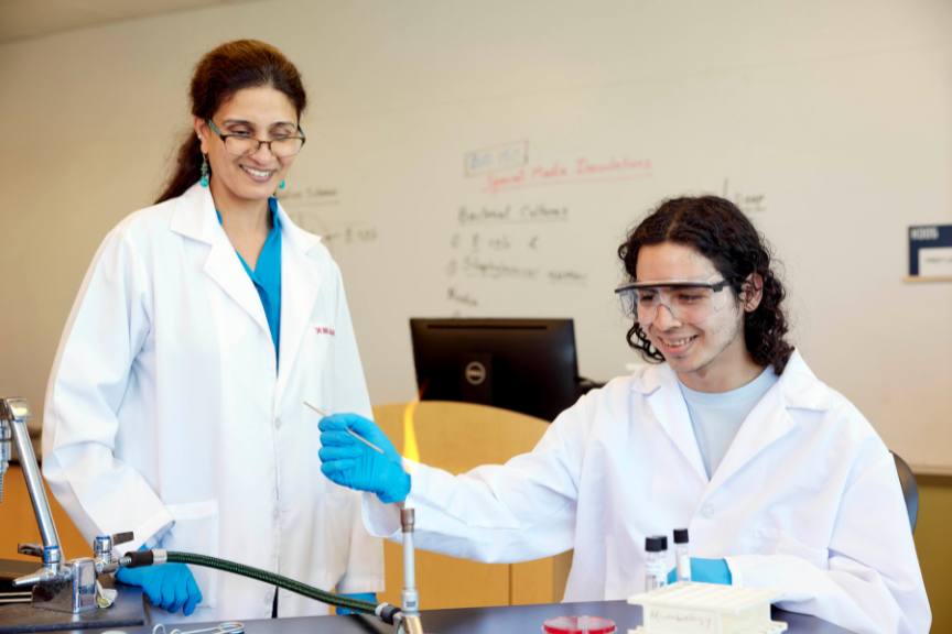 Two scientists in lab coats and safety glasses work at a laboratory bench. One is holding a test tube near a Bunsen burner flame while the other observes, both smiling. Lab equipment and a whiteboard are in the background.