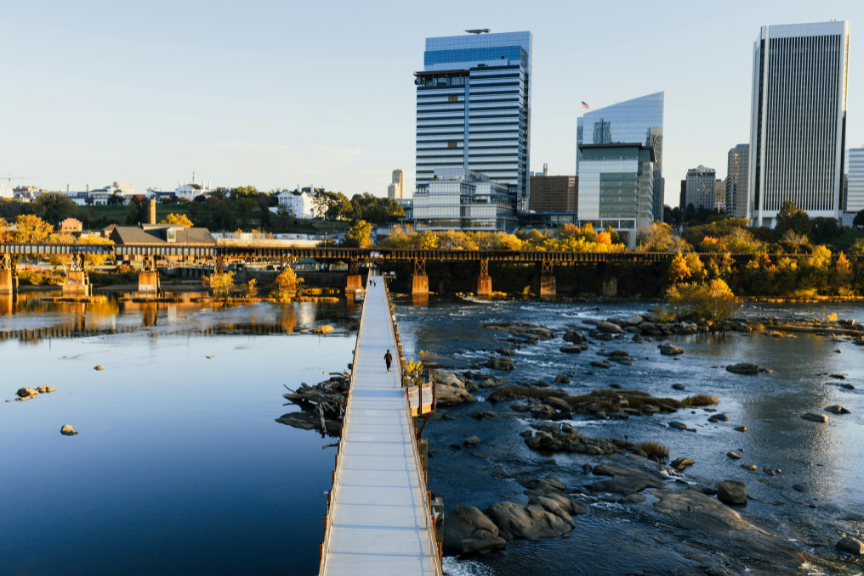A long pedestrian bridge crosses a river toward a city skyline with modern high-rise buildings and autumn trees lining the riverbank under a clear sky.