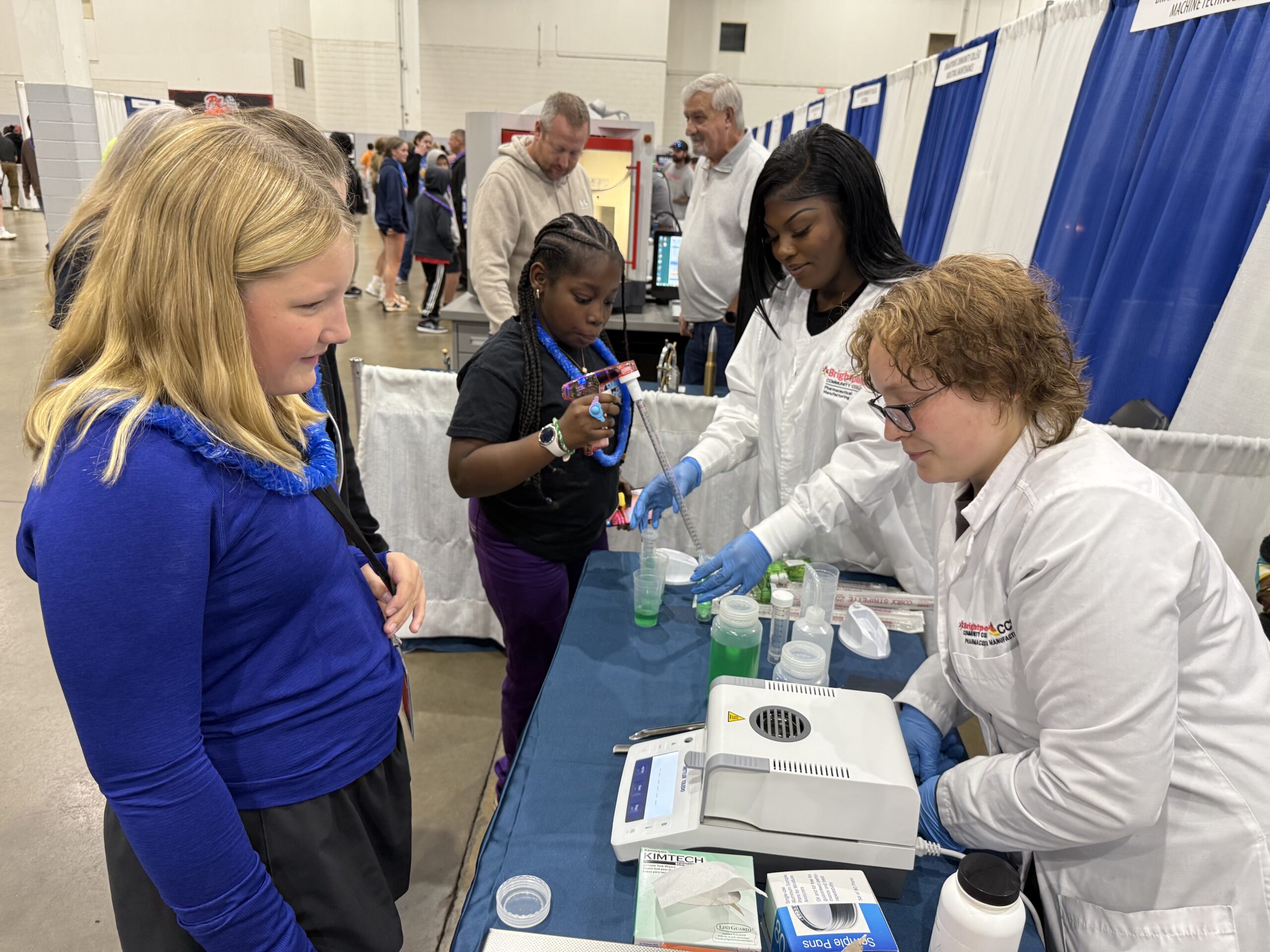 A young girl watches as two women in lab coats conduct a science experiment with a child at a booth, while people walk in the background at an indoor event.