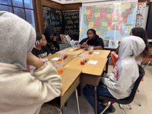 Six students sit around a table in a classroom, talking with a teacher. Colored drinks, science materials, and a U.S. map are on the wall behind them. The group appears to be discussing an experiment.