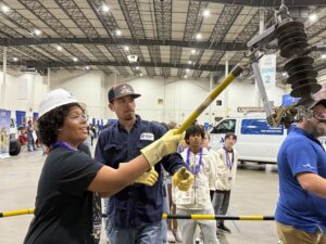 A woman in a hard hat and gloves uses a long tool to handle equipment, guided by a man in a cap and work shirt, while others observe in a large indoor industrial space.