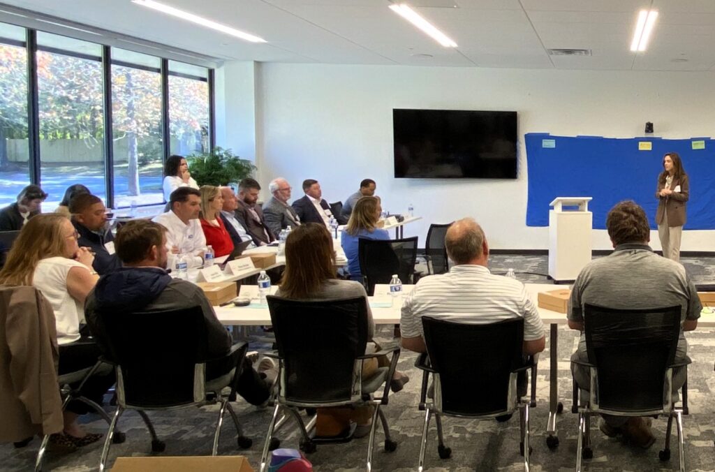 A woman stands and speaks at a podium in a conference room, addressing members of the Construction Sector Partnership. Daylight fills the room from the large windows behind the participants on the left.