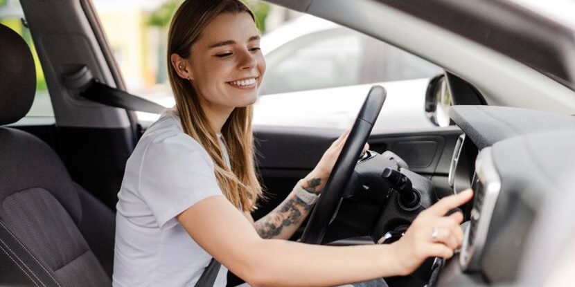 A young woman with long hair and a tattoo on her arm smiles while adjusting the car stereo, sitting in the drivers seat with her seatbelt fastened.
